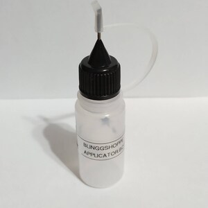 May include: A clear plastic applicator bottle with a black cap and a fine-tipped nozzle. The bottle has a label that reads "BLINGGSHOPPE APPLICATOR BOTTLE". The nozzle is designed for precise application of liquids.