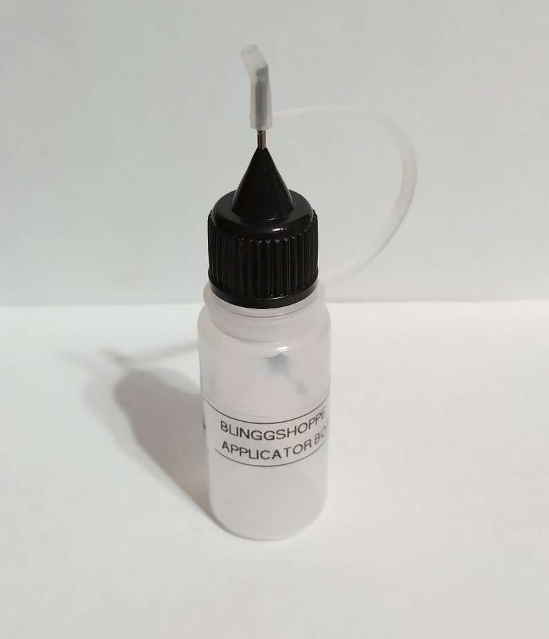 May include: A clear plastic applicator bottle with a black cap and a fine-tipped nozzle. The bottle has a label that reads "BLINGGSHOPPE APPLICATOR BOTTLE". The nozzle is designed for precise application of liquids.