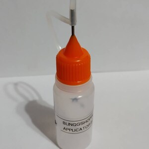 May include: A small, clear plastic bottle with an orange cap and a needle applicator. The bottle has a label with the text "BLINGGSHOP APPLICATOR B". The needle is attached to a clear plastic tube.