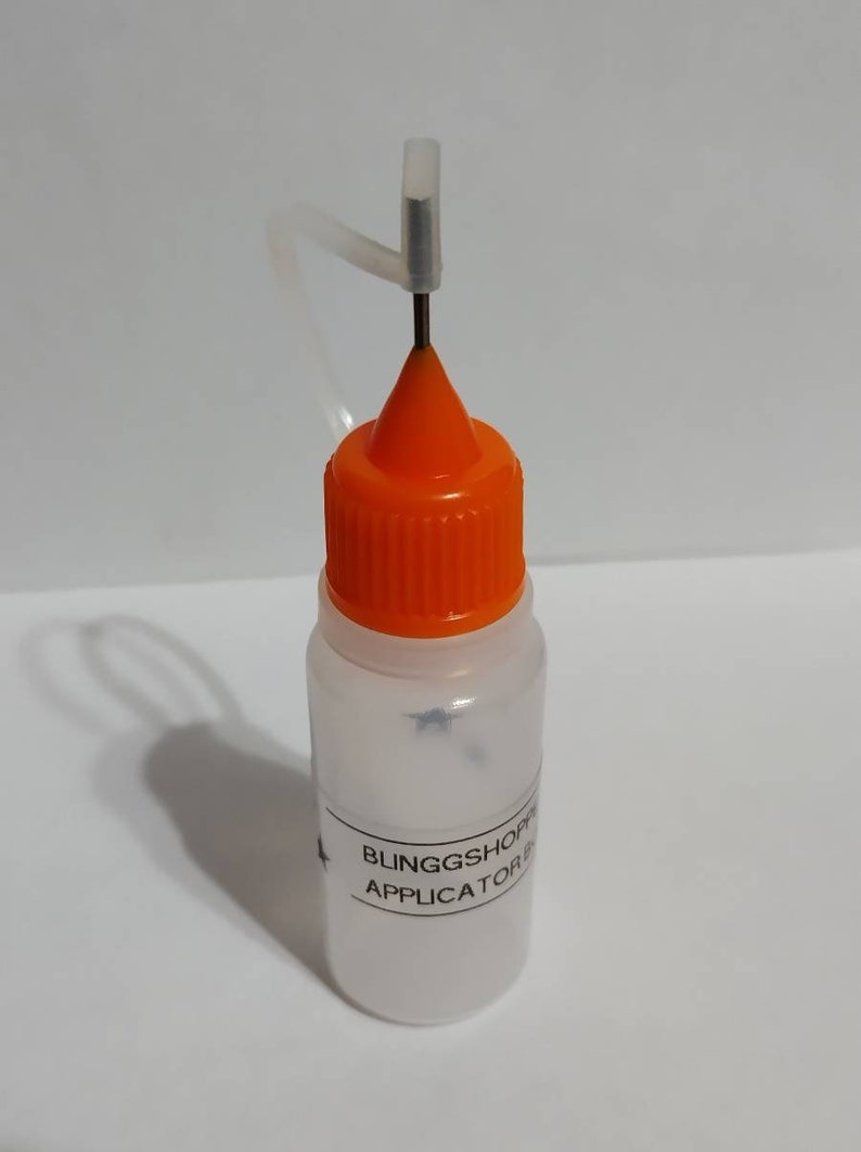 May include: A small, clear plastic bottle with an orange cap and a needle applicator. The bottle has a label with the text "BLINGGSHOP APPLICATOR B". The needle is attached to a clear plastic tube.