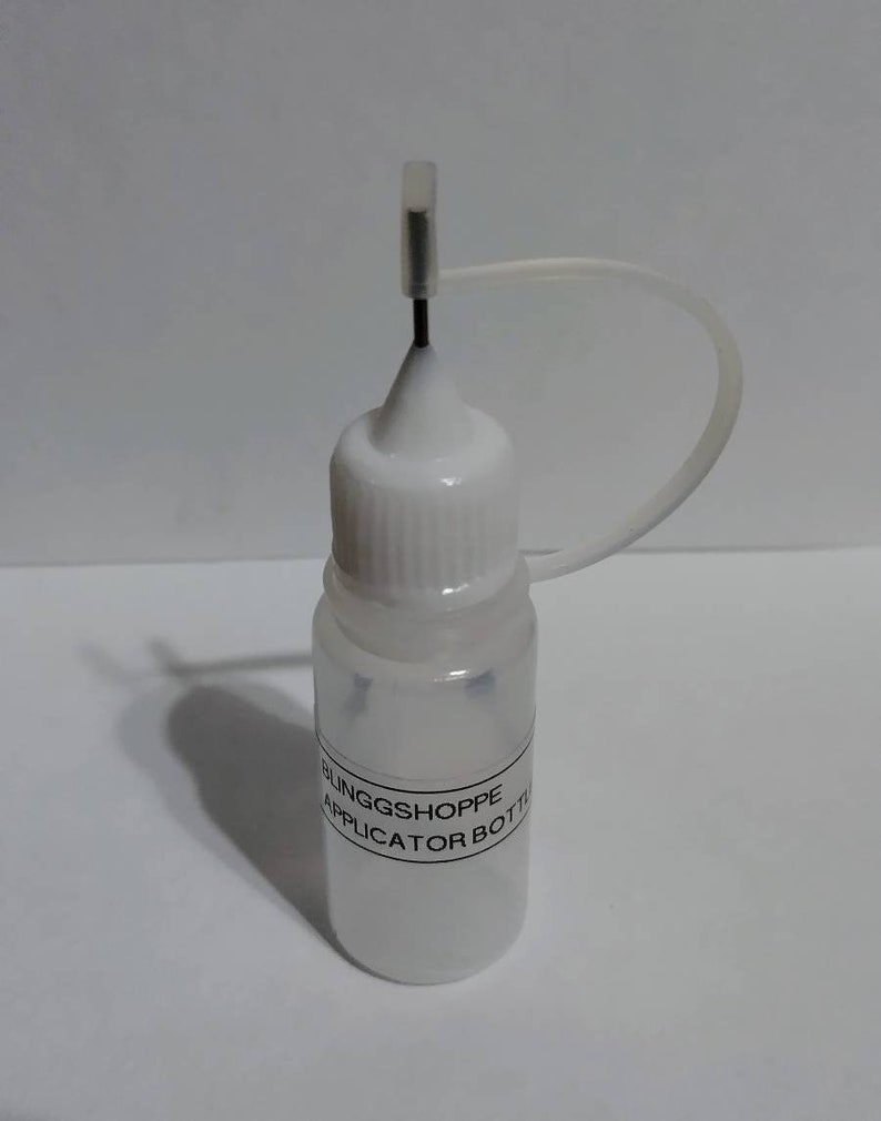 May include: A small, clear plastic applicator bottle with a white cap and a thin, black needle. The bottle has a label that reads "BLINGGSHOPPE APPLICATOR BOTTLE". A clear plastic tube is attached to the cap.