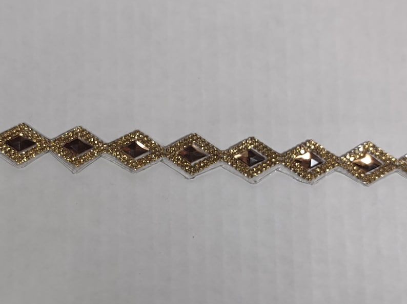 May include: A gold trim with a row of diamond-shaped rhinestones. The rhinestones have a clear center and a gold border.