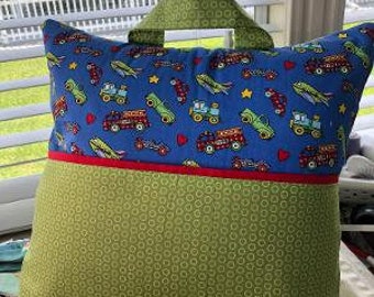 Pode incluir: Uma almofada verde e azul com uma alça. A almofada é coberta por um tecido com um padrão de caminhões de brinquedo vermelhos, amarelos e azuis. A parte de trás da almofada é verde sólida.