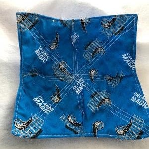 May include: A blue square bowl cozy with an Orlando Magic basketball team design. The design includes the team name and logo in white. The cozy is made of fabric and is designed to be used in the microwave.