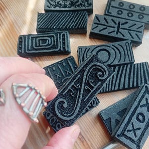 May include: A collection of dark gray rectangular stamps with various embossed patterns, including geometric shapes and stylized designs. The stamps are arranged on a light-colored surface, with one held by a hand. A silver ring with a fan-like design is also visible.