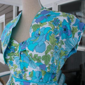 May include: A sleeveless dress featuring a vibrant floral pattern in shades of bright blue, light blue, purple, and lime green on a white background. The bodice has a sweetheart neckline and a wide, folded collar detail that extends over the shoulders.