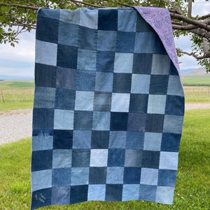 May include: A patchwork quilt constructed from square denim fabric pieces in various shades of blue, arranged in a grid pattern. The reverse side of the quilt features a light purple fabric with a dark purple paisley print pattern throughout.