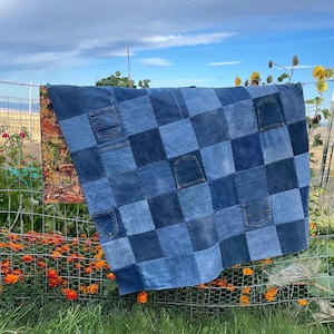 May include: A patchwork quilt constructed from various shades of blue denim fabric squares, featuring several integrated denim pockets. The reverse side of the quilt displays a printed fabric pattern showing deer in an autumn forest setting.