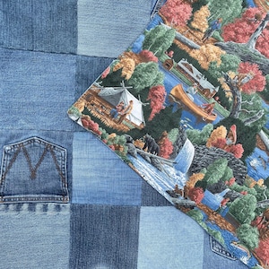 May include: A patchwork textile featuring a combination of blue denim squares with a visible pocket and a printed fabric section depicting a colorful outdoor wilderness scene with a tent, a canoe on a river, trees, and a waterfall.