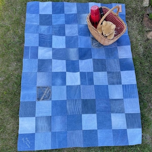 May include: A rectangular patchwork quilt constructed from various shades of blue denim fabric squares. A woven wicker basket sits on the quilt, containing a red thermos, a black pen, and a small tan teddy bear. One denim pocket is visible.