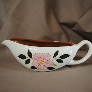Puede incluir: Salsera de cerámica blanca con interior y asa marrones. El exterior presenta un diseño floral rosa con hojas verdes. La salsera está diseñada con un pico para verter fácilmente.