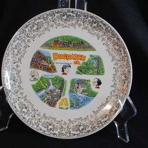 May include: A decorative plate with a gold filigree border and a central image featuring the text "Dogpatch USA" and cartoon illustrations. The plate is white with gold accents and is displayed on a clear stand.