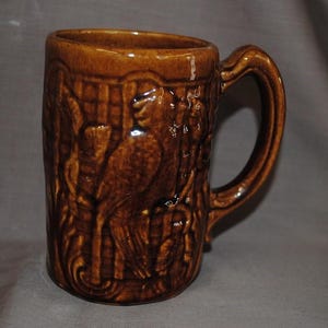 May include: A brown ceramic mug with a handle. The mug features a detailed relief design of a bird and foliage. The mug has a glossy finish and is suitable for hot or cold beverages. The mug is approximately 5 inches tall.