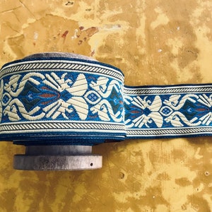 May include: A roll of decorative trim featuring a repeating pattern of stylized floral and geometric shapes in cream, blue, and teal. The trim is wound on a wooden spool, showcasing its intricate design and color palette.