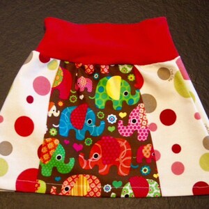 May include: A white skirt with red, pink, and gray polka dots. The skirt has a brown panel with colorful elephants and flowers. The skirt has a red waistband.