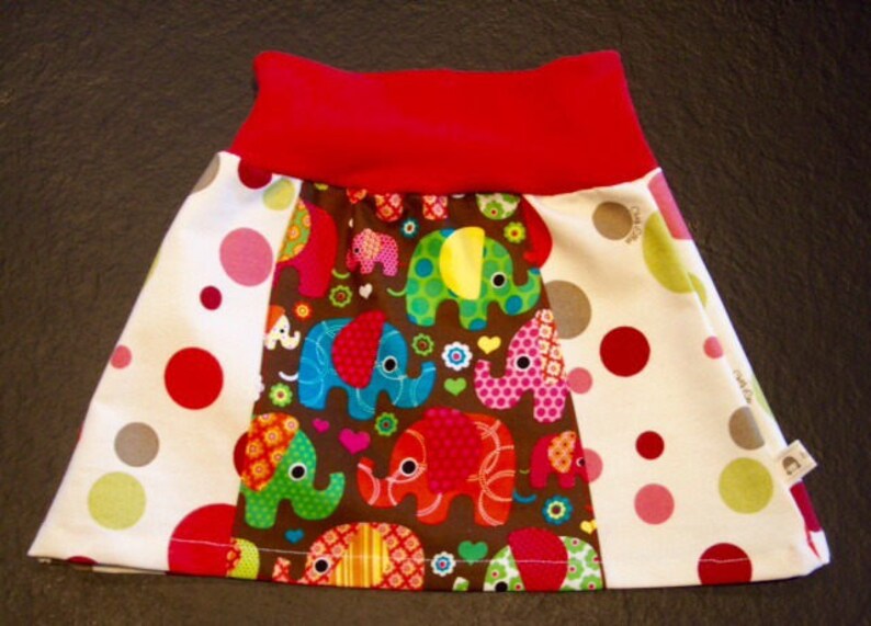 May include: A white skirt with red, pink, and gray polka dots. The skirt has a brown panel with colorful elephants and flowers. The skirt has a red waistband.