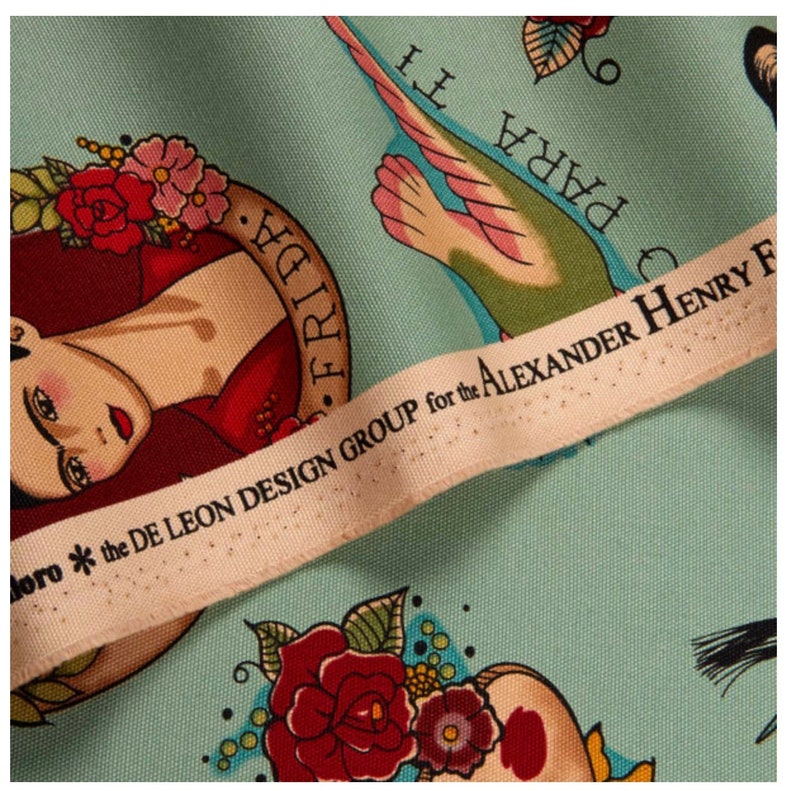 May include: Close-up of a fabric with a light blue background featuring a Frida Kahlo illustration, roses, and a bird. The fabric includes the text "FRIDA" and "PARA TI". A cream-colored label reads "the DE LEON DESIGN GROUP for the ALEXANDER HENRY F".