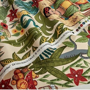 May include: A cream-colored fabric with a colorful tropical print. The print features a woman with flowers in her hair, a snake, and various tropical plants. The fabric has a label that reads "De Leon Design Group * The Alexander Fabrics Collection".