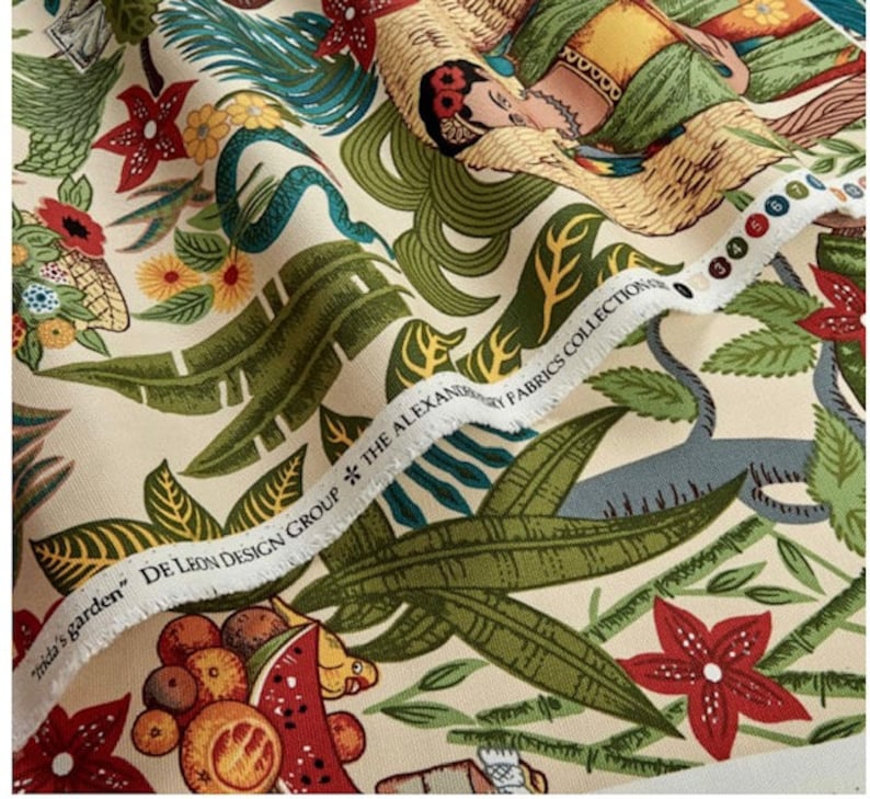 May include: A cream-colored fabric with a colorful tropical print. The print features a woman with flowers in her hair, a snake, and various tropical plants. The fabric has a label that reads "De Leon Design Group * The Alexander Fabrics Collection".