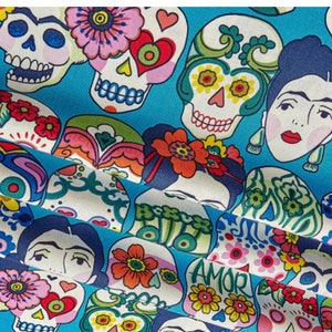 May include: A blue fabric with a repeating pattern of colorful sugar skulls and floral designs. The skulls have black eyes and are decorated with flowers and hearts. The fabric also features images of women with flowers in their hair.
