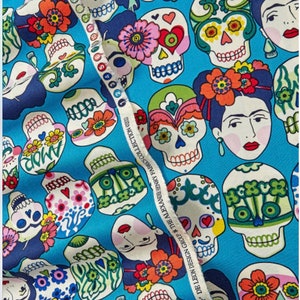 May include: A blue fabric with a repeating pattern of colorful sugar skulls and Frida Kahlo portraits. The fabric has the text "DE LEON DESIGN GROUP THE ALEXANDER HENRY FABRES COLLECTION 2023" printed on the side.
