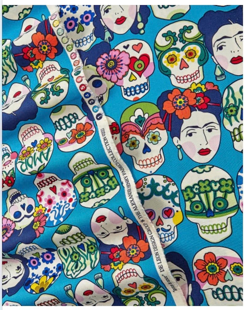 May include: A blue fabric with a repeating pattern of colorful sugar skulls and Frida Kahlo portraits. The fabric has the text "DE LEON DESIGN GROUP THE ALEXANDER HENRY FABRES COLLECTION 2023" printed on the side.