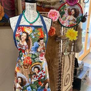May include: A colorful apron with a blue denim trim features a vintage-inspired design with images of women wearing sombreros and flowers. The apron is displayed on a mannequin with a turquoise necklace.