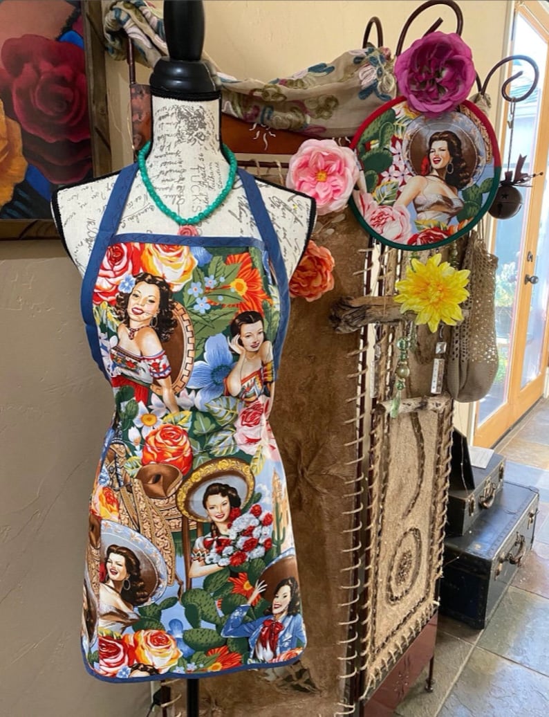 May include: A colorful apron with a blue denim trim features a vintage-inspired design with images of women wearing sombreros and flowers. The apron is displayed on a mannequin with a turquoise necklace.