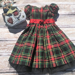 May include: A black, red, and green plaid dress with a red ribbon and bow. The dress has a black tulle underskirt.