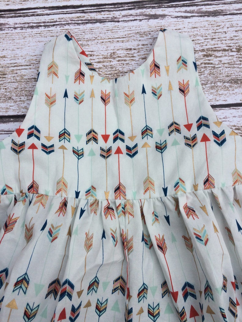 May include: A white sleeveless dress with a pattern of colorful arrows. The dress has a gathered skirt and a simple neckline.