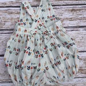 May include: A white baby onesie with a criss-cross back and a bow. The onesie has a pattern of colorful arrows on a light blue background.