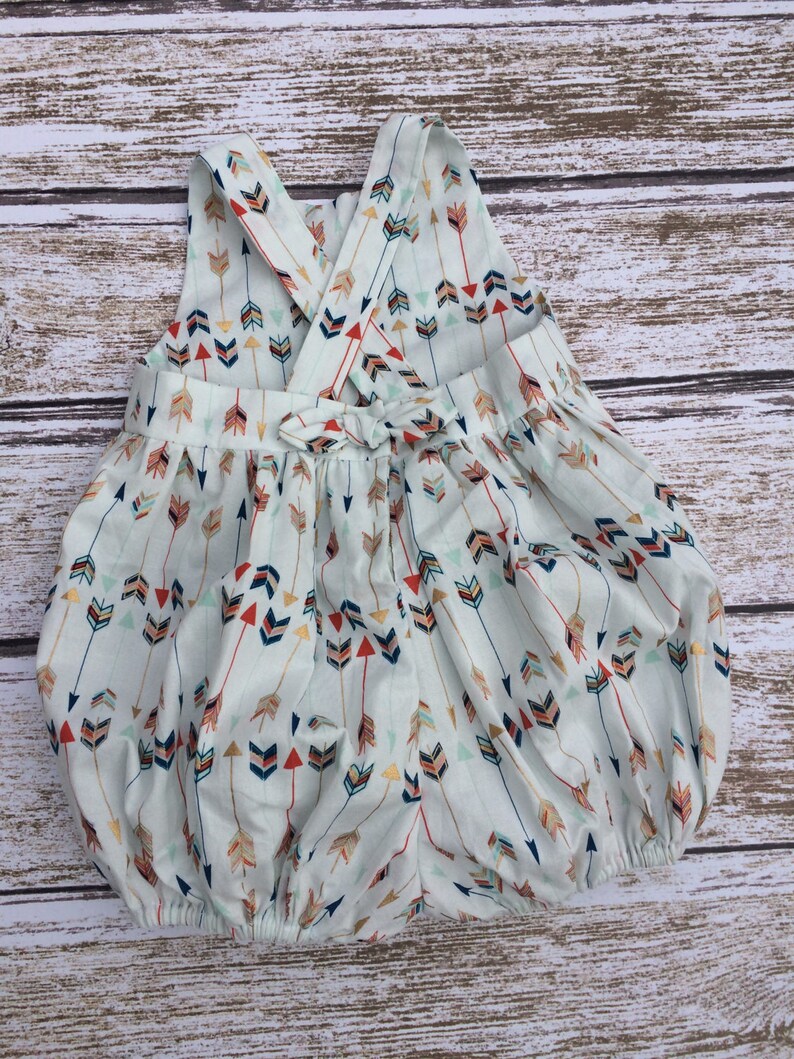 May include: A white baby onesie with a criss-cross back and a bow. The onesie has a pattern of colorful arrows on a light blue background.