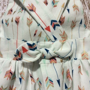May include: A white sleeveless dress with a criss-cross strap design and a bow at the waist. The dress is covered in a pattern of colorful arrows with gold accents.