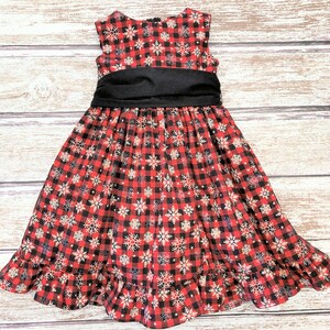 May include: A red and black plaid dress with a snowflake pattern and a black satin sash. The dress has a ruffled hem.