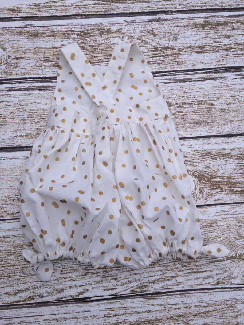 May include: White baby onesie with gold polka dots and a criss-cross strap back.