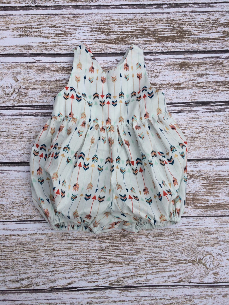 May include: A white baby onesie with a pattern of colorful arrows. The onesie has a sleeveless design and a gathered skirt.
