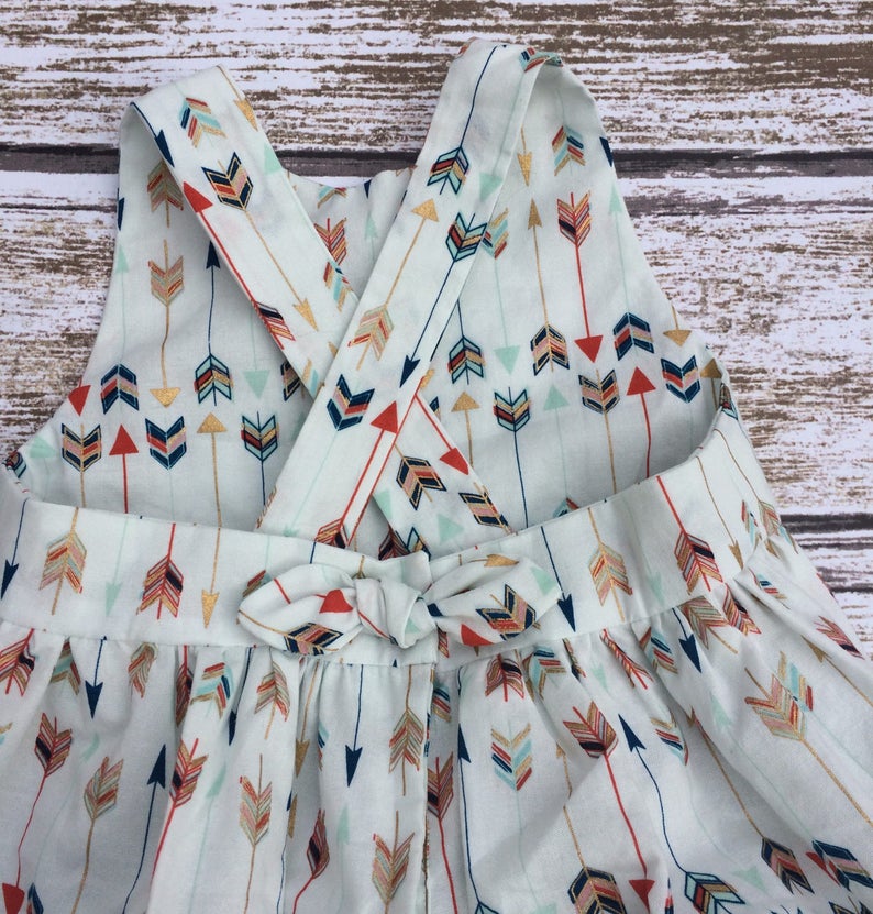 May include: A white sleeveless dress with a criss-cross back and a bow tie at the waist. The dress is covered in a pattern of colorful arrows.
