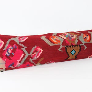 Puede incluir: Un cojín largo y rectangular con un fondo rojo oscuro y un estampado floral tejido. El diseño presenta flores rosas, grises y naranjas, junto con una forma de jarrón marrón con un diamante azul. El cojín es un elemento decorativo para el hogar.