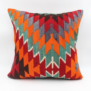 Puede incluir: Una almohada de tiro colorida con un patrón geométrico en tonos de rojo, naranja, verde, azul y marrón. La almohada está hecha de tejido tejido y tiene una forma cuadrada.