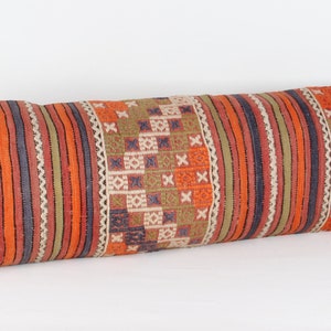 Puede incluir: Una almohada larga y rectangular con un patrón geométrico colorido. La almohada está hecha de una tela tejida con rayas y cuadrados en tonos naranja, rojo, verde y marrón.