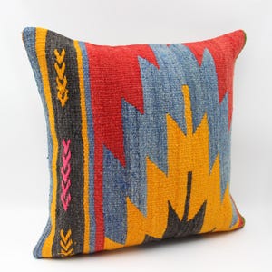 May include: Square cushion with a geometric design. The cushion has a central pattern in yellow, blue, and black, framed by a red border. The side of the cushion has a vertical stripe with yellow and pink accents.