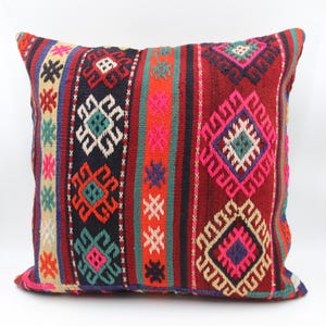 Pode incluir: Uma almofada decorativa quadrada com um padrão geométrico tecido. A almofada apresenta um fundo vermelho com listras verticais em várias cores, incluindo preto, laranja, rosa e verde-azulado. O design inclui formas de diamante e cruz.