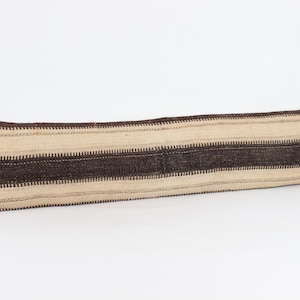 May include: A long, rectangular lumbar cushion featuring a woven pattern of horizontal stripes in alternating shades of dark brown and cream. The fabric has a textured, coarse appearance with visible stitching along the edges of the cushion cover.