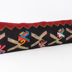 Puede incluir: Un cojín largo y rectangular con fondo negro y diseños bordados coloridos. El borde superior es rojo oscuro con un patrón en zigzag. El cojín presenta motivos geométricos y florales en varios colores.