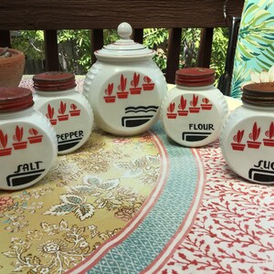 May include: A set of five white milk glass kitchen canisters with red metal lids. The containers feature red potted plant graphics and black text labels reading Salt, Pepper, Flour, and Sugar. The set includes four round jars and one larger lidded jar.