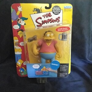 May include: A plastic Barney figure from The Simpsons, with a pink shirt, blue pants, and a yellow head. The figure is holding a beer mug and has a voice activation feature. The packaging includes the text "Intelli-Tronic Voice Activation" and "Barney".