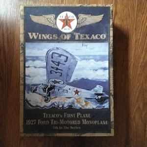 May include: A vintage box featuring a painting of a silver airplane with the number 3443 on the side. The box is labeled "Wings of Texaco" and "Texaco's First Plane 1927 Ford Tri-Motored Monoplane 7th in the Series".