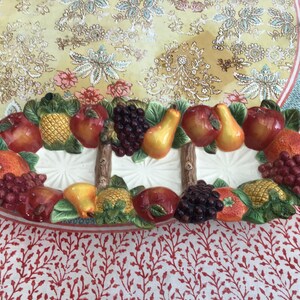 May include: An elongated ceramic serving tray featuring a three-dimensional relief of assorted fruits including red apples, yellow pears, purple grapes, pineapples, and oranges, separated by two brown textured branch-like dividers on a white base.