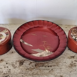 May include: Three-piece set of lacquered Japanese serving pieces. The set includes two lidded containers and a large, round tray. The tray and containers are red with decorative designs. The tray features a landscape scene.
