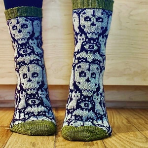 May include: Knitted socks featuring a repeating skull design in white and navy blue. The socks have green cuffs, heels, and toes. The socks are worn over black leggings.